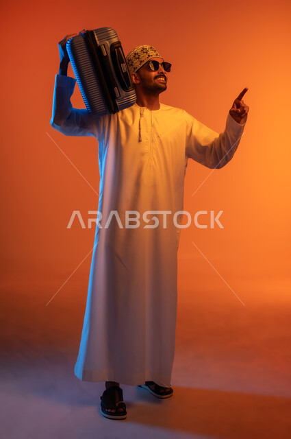 The concept of tourism and travel, preparing for the trip, having a good time on vacation, a portrait of a young Arab Gulf Omani man wearing a dishdasha and a turban, carrying a travel bag on his shoulder, pointing with his index finger at something, colored background