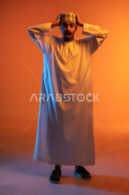 المفاجأة و الصدمة عند رؤية شيء ما، الانزعاج عند سماع خبر سيء, الوقوع في مصيبة, النظر الى الكاميرا بتعابير التعجب, بورتريه لشاب عربي خليجي عماني يرتدي الدشداشة والكمه يضع يديه على رأس بتعابير الدهشة و الذهول، الجسم بالطول الكامل, خلفيه ملونة