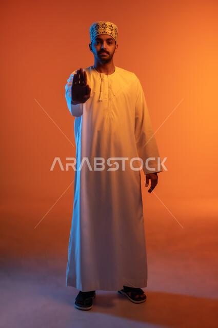 الوقوف بغضب ورفض أمر محدد, الصمت والتوقف عن الكلام, طلب عدم التدخل وخفض الصوت والتهديد, الامتناع عن فعل أمر معين, بوتريه لشاب عربي خليجي عماني يرتدي الكمه و الدشداشة يرفع كفه للأعلى بإيماءات الانزعاج, الجسم بالطول الكامل, خلفية ملونة