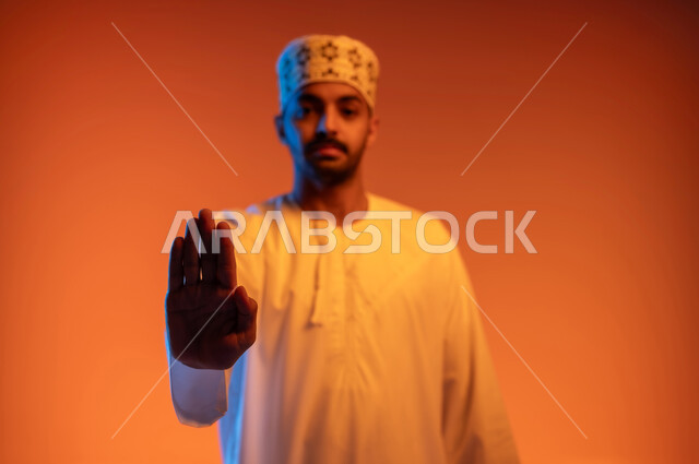 طلب عدم التدخل وخفض الصوت والتهديد, الوقوف بغضب ورفض أمر محدد, الصمت والتوقف عن الكلام, الامتناع عن فعل أمر معين, بوتريه مقرب لشاب عربي خليجي عماني يرتدي الكمه و الدشداشة يرفع كفه للأعلى بإيماءات الانزعاج, خلفية ملونة