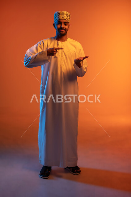 الاشارة بإصبع السبابة إلى شيء محدد, رفع وحمل عنصر معين, الوقوف والنظر إلى الكاميرا بإيماءات السرور, بورتريه لشاب عربي خليجي عماني مبتسم يرتدي الكمه و الدشداشة يحمل بيده شيء ما, الطول الكامل للجسم, خلفية ملونة