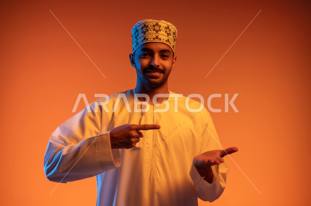رفع وحمل عنصر معين, الوقوف والنظر إلى الكاميرا بإيماءات السرور, الاشارة بإصبع السبابة إلى شيء محدد, بورتريه مقرب لشاب عربي خليجي عماني مبتسم يرتدي الكمه و الدشداشة يحمل بيده شيء ما, خلفية ملونة
