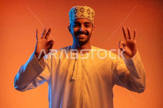تعابير التميز والاعجاب, بورتريه مقرب لشاب عربي خليجي عماني يرتدي الدشداشة والكمه يرفع يديه للاعلى بإيماءات القبول و الموافقة والرضا, علامة الجودة والإتقان, خلفية ملونة