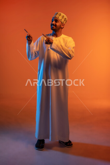 توضيح و تحديد موقع معين, الوقوف باستقامة والنظر إلى شيء ما بتعابير السرور, بورتريه لشاب عربي خليجي عماني مبتسم يرتدي الكمه و الدشداشة يرفع اصابع السبابة يشير إلى هدف محدد, الطول الكامل للجسم, خلفية ملونة