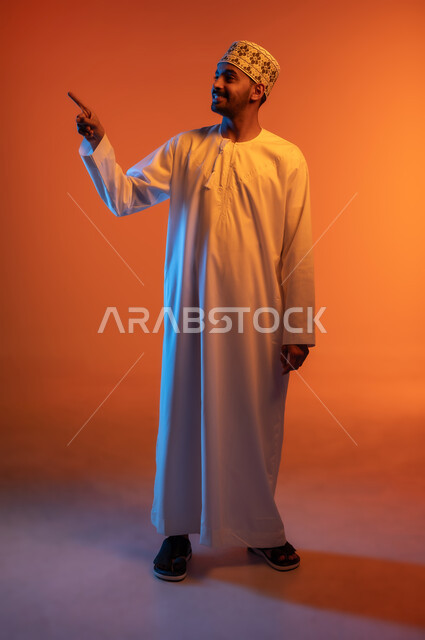 الوقوف باستقامة والنظر إلى شيء ما بتعابير السرور, الإشارة إلى هدف محدد, بورتريه لشاب عربي خليجي عماني مبتسم يرتدي الكمه و الدشداشة يرفع اصبع السبابة لتحديد وتوضيح موقع معين, الجسم بالطول الكامل, خلفية ملونة