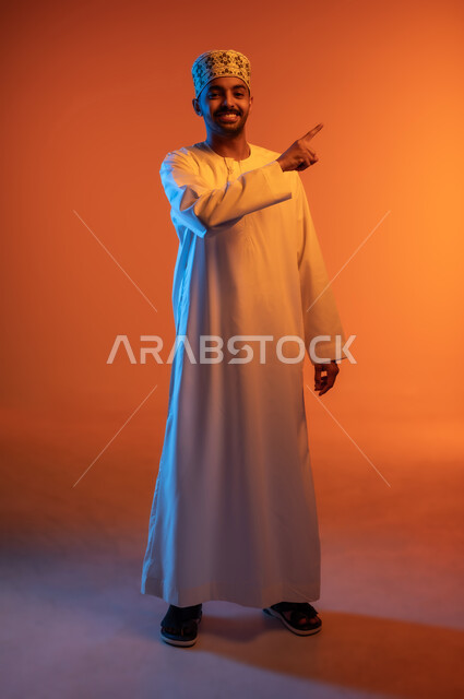 الإشارة إلى هدف محدد, الوقوف باستقامة والنظر إلى الكاميرا بتعابير السرور, بورتريه لشاب عربي خليجي عماني مبتسم يرتدي الكمه و الدشداشة يرفع اصبع السبابة لتحديد وتوضيح موقع معين, الجسم بالطول الكامل, خلفية ملونة