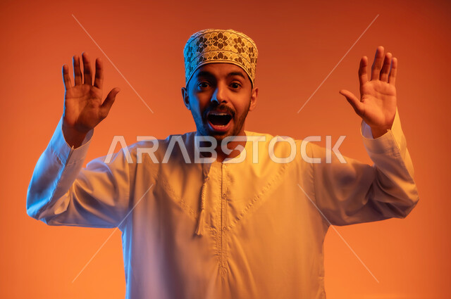 النظر الى الكاميرا بايماءات المفاجأة ,بورتريه مقرب لشاب عربي خليجي عماني يرتدي الدشداشة و الكمه يرفع يديه الى للاعلى بإيماءات الاستسلام، الشعور بالذهول والصدمه, خلفيه ملونة