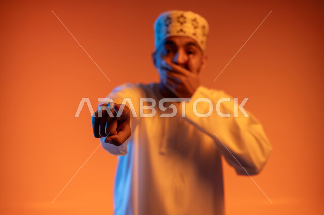 الاشارة باصبع السبابة الى شيء ما بحركات الاستهزاء، بورتريه مقرب لرجل عربي خليجي عماني يرتدي الكمه و الدشداشة يضع يده على فمه بإيماءات الضحك الشديد، النظر الى الكاميرا بتعابير السخرية, خلفية ملونة