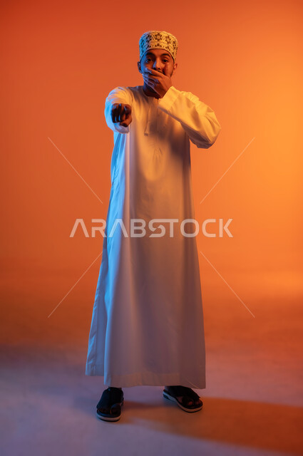 النظر الى الكاميرا بتعابير السخرية, الاشارة باصبع السبابة الى شيء ما بحركات الاستهزاء، بورتريه لرجل عربي خليجي عماني يرتدي الكمه و الدشداشة يضع يده على فمه بإيماءات الضحك الشديد، الجسم بالطول الكامل، خلفية ملونة