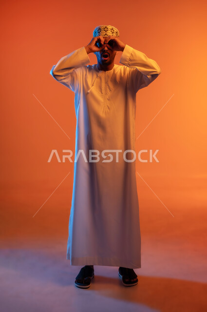 الرؤية المستقبلية و التخطيط للأهداف والطموحات, حب المعرفة والاستكشاف والمغامرة, بورتريه لشاب عربي خليجي عماني مبتسم يرتدي الكمه و الدشداشة يضع يده على عينه على شكل عدسة للنظر عن قرب, صورة الجسم بالطول الكامل, خلفية ملونة