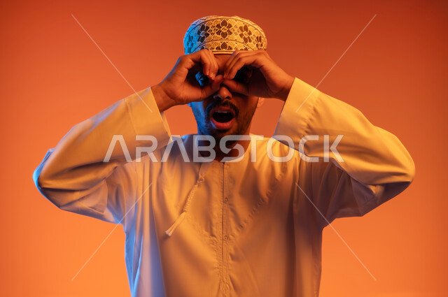 النظر الى شيء ما بتعابير الاندهاش والاستغراب, التخطيط للأهداف والطموحات المستقبلية, رؤية الأشياء البعيدة, بورتريه مقرب لشاب عربي خليجي عماني يرتدي الكمه والدشداشة يضع يديه على عينيه بشكل عدسة, حب الاكتشاف والمعرفة والمغامرة, خلفية ملونة
