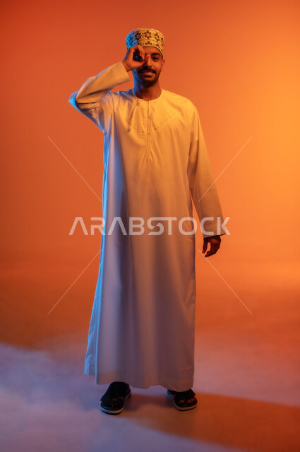 حب المعرفة والاستكشاف والمغامرة, التخطيط والرؤية المستقبلية للأهداف والطموحات, بورتريه لشاب عربي خليجي عماني مبتسم يرتدي الكمه و الدشداشة يضع يده على عينه على شكل عدسة للنظر عن قرب, صورة الجسم بالطول الكامل, خلفية ملونة
