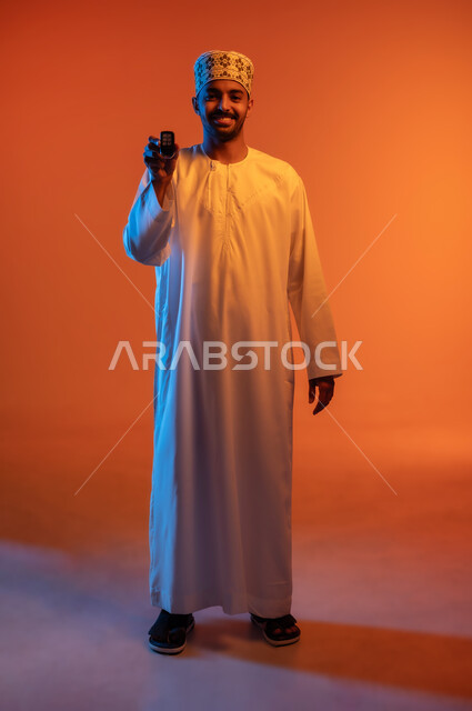 مفهوم القياده في سلطنة عمان, النظر الى الكاميرا بتعابير السرور, ايماءات السرور بامتلاك سيارة جديده, بورتريه لرجل عربي خليجي عماني مبتسم يرتدي الكمه و الدشداشة يحمل بيده مفتاح التحكم عن بعد, الجسم بالطول الكامل, خلفيه ملونة