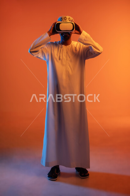استخدام التقنيات والأجهزة المتطورة, الاندماج و التفاعل ومحاكاة العالم الخيالي, بورتريه لشاب عربي خليجي عماني يرتدي الكمه و الدشداشة يمسك بيده نظارة الواقع الافتراضي, التسلية ومشاهدة الأفلام في وقت الفراغ, الجسم بالطول الكامل, خلفية ملونة