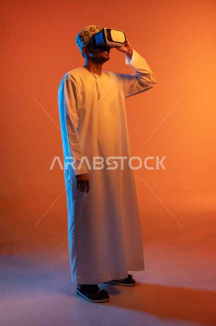 الاندماج و التفاعل ومحاكاة العالم الخيالي, بورتريه لشاب عربي خليجي عماني يرتدي الكمه و الدشداشة يمسك بيده نظارة الواقع الافتراضي, التسلية ومشاهدة الأفلام في وقت الفراغ, استخدام التقنيات والأجهزة المتطورة, الجسم بالطول الكامل, خلفية ملونة