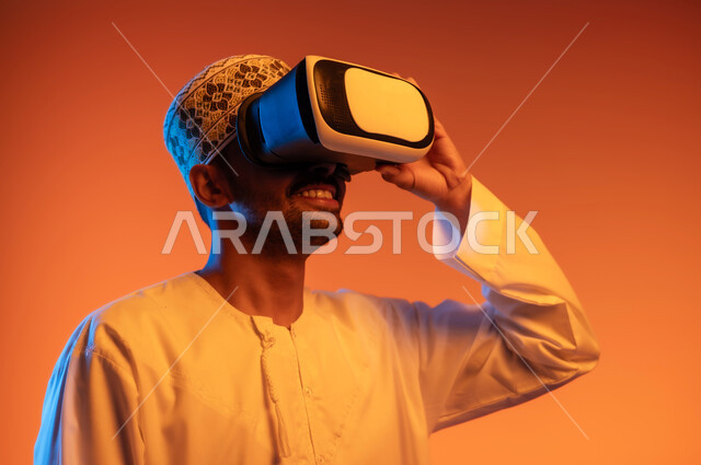 التسلية ومشاهدة الأفلام في وقت الفراغ, استخدام التقنيات والأجهزة المتطورة, الاندماج و التفاعل ومحاكاة العالم الخيالي, بورتريه مقرب لشاب عربي خليجي عماني يرتدي الكمه و الدشداشة يمسك بيده نظارة الواقع الافتراضي, خلفية ملونة