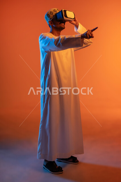 الاندماج ومحاكاة العالم الخيالي, بورتريه لشاب عربي خليجي عماني يرتدي الكمه و الدشداشة يمسك بيده نظارة الواقع الافتراضي يشير باصبع السبابة الى شيء ما, التسلية ومشاهدة الأفلام في وقت الفراغ, استخدام التقنيات والأجهزة المتطورة, الجسم بالطول الكامل, خلفية ملونة