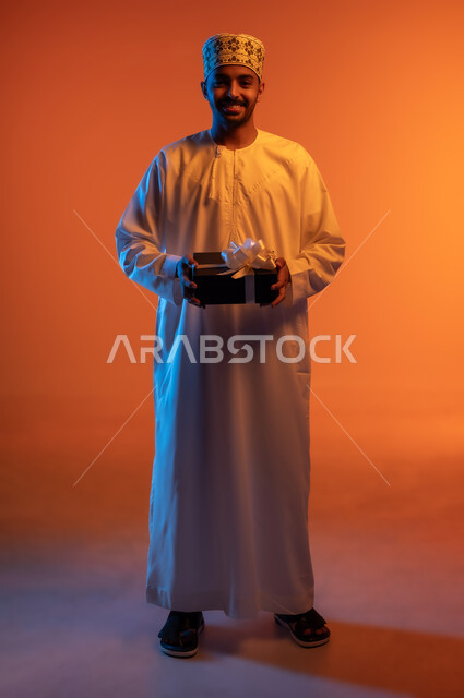 تقديم الهدايا بالأعياد والمناسبات الخاصة، التعبير عن الحب والتقدير والاحترام، مفهوم الشكر والامتنان، النظر الى الكاميرا بتعابير السرور, بورتريه لرجل عربي عماني خليجي يرتدي الكمه و الدشداشة يحمل بيديه صندوق هدية مغلف, الجسم بالطول الكامل، خلفية ملونة