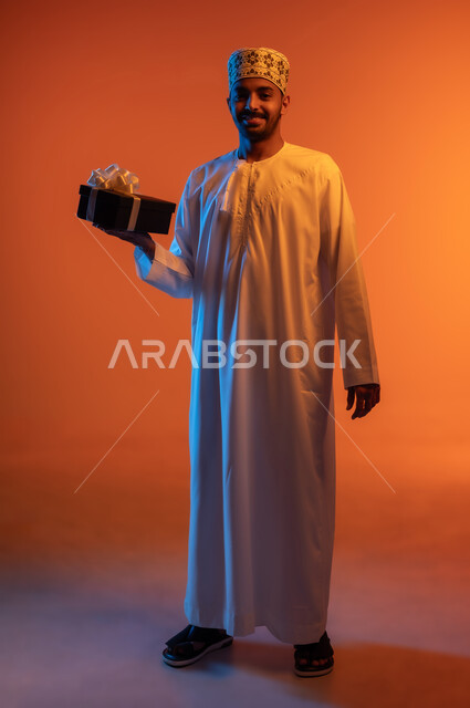 مفهوم الشكر والامتنان، تقديم الهدايا بالأعياد والمناسبات الخاصة، التعبير عن الحب والتقدير والاحترام، النظر الى الكاميرا بتعابير السرور, بورتريه لرجل عربي عماني خليجي يرتدي الكمه و الدشداشة يحمل بيده صندوق هدية مغلف, الجسم بالطول الكامل، خلفية ملونة