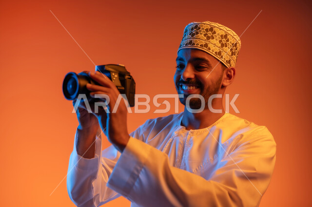 Using high-resolution digital cameras, Omani jobs and professions, professional photography, close-up portrait of a smiling professional Arab Gulf Omani photographer wearing a turban and a dishdasha taking souvenir photos with a camera, colored background