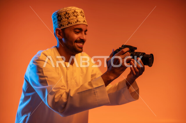 احتراف فن التصوير الفوتوجرافي باستخدام كاميرات رقمية عالية الدقة, وظائف ومهن عمانية, بورتريه مقرب من الجانب لمصور محترف عربي خليجي عماني يرتدي الكمه و الدشداشة يقوم بالتقاط الصور التذكارية عبر كاميرا فوتوجرافية, خلفية ملونة