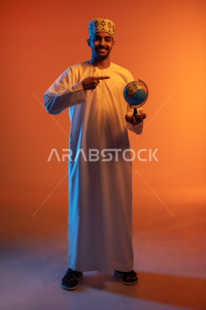 The concept of love of research and exploration of continents in geography, pointing to the globe, close-up portrait of a smiling Omani Gulf Arab man wearing a turban and a dishdasha pointing with his index finger to the world map, expressions of pleasure and joy at belonging to the Arab countries, colored background