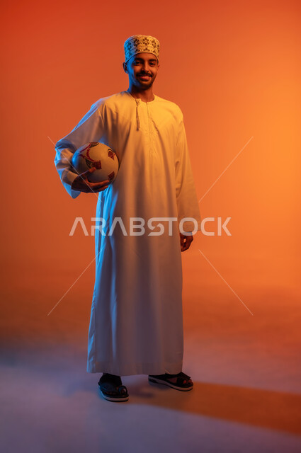 Practicing favorite hobbies, standing straight and looking at the camera, portrait of an Arab Omani Gulf player wearing a turban and a dishdasha holding a football under his arm preparing to start the match, entertainment and recreation with group sports, full body image, colored background