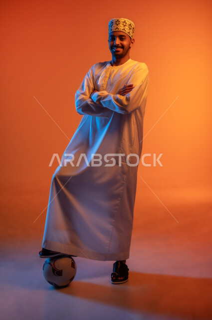 Standing straight with arms crossed, professional football player, looking at camera with confident gestures, portrait of an Arab Omani Gulf player wearing a kimono and a dishdasha preparing to start the match, entertainment and recreation with group sports, full body image, colorful background