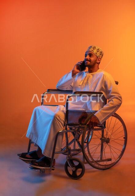 Making a voice call via mobile, caring for the psychology of people with disabilities and giving them positive energy, communicating and chatting with relatives, laughing and chatting with friends, portrait of a disabled Gulf Arab Omani man wearing a turban and a dishdasha sitting in a wheelchair and holding a mobile phone in his hand, colored background