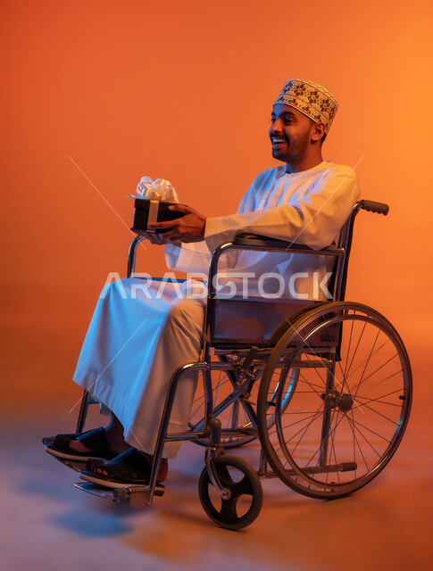 اهتمام ودعم المجتمع العماني لذوي الهمم, مناسبات و أعياد سعيدة، بورتريه لشاب عربي خليجي عماني مقعد يرتدي الكمه و الدشداشة يجلس على كرسي متحرك يحمل بيديه علب الهدايا المغلفة، النظر الى شيء ما بإيماءات الدهشة والفرح, شعور المعاقين بالحب و التقدير، خلفية ملونة