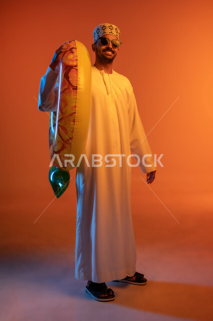 قضاء عطلة صيفيه ممتعه ،رحلة سياحية ترفيهيه، النظر الى الكاميرا بايماءات السرور, بورتريه لشاب عربي خليجي عماني يرتدي الدشداشة والكمه والنظارات الشمسية يحمل على كتفه عوامة السباحة، مفهوم التسلية في أوقات الفراغ، صورة الجسم بالطول الكامل, خلفية ملونة