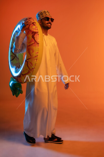 ايماءات المشي والذهاب لقضاء عطلة صيفيه ممتعه ،رحلة سياحية ترفيهيه، بورتريه لشاب عربي خليجي عماني يرتدي الدشداشة والكمه والنظارات الشمسية يحمل على كتفه عوامة السباحة، مفهوم التسلية في أوقات الفراغ، صورة الجسم بالطول الكامل, خلفية ملونة