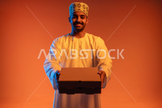 Delivering orders on time and in good condition, close-up portrait of an Arab Gulf Omani man wearing a kimono and a dishdasha holding a closed cardboard box, receiving purchases and delivering goods to customers, looking at the camera with an expression of pleasure, colorful background