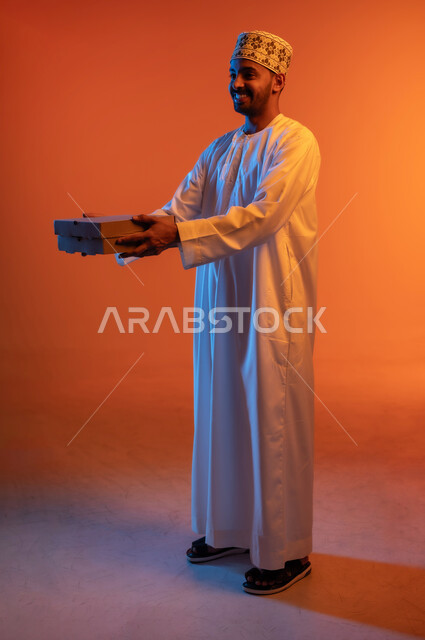 توصيل الطلبيات بالوقت المحدد و بشكل سليم, بورتريه من الجانب لرجل عربي خليجي عماني يرتدي الكمه و الدشداشة يحمل بيده صندوق كرتوني مغلق, إستلام المشتريات و تسليم البضائع للزبائن, النظر الى شيء ما بتعابير السرور, صورة الجسم بالطول الكامل، خلفية ملونة