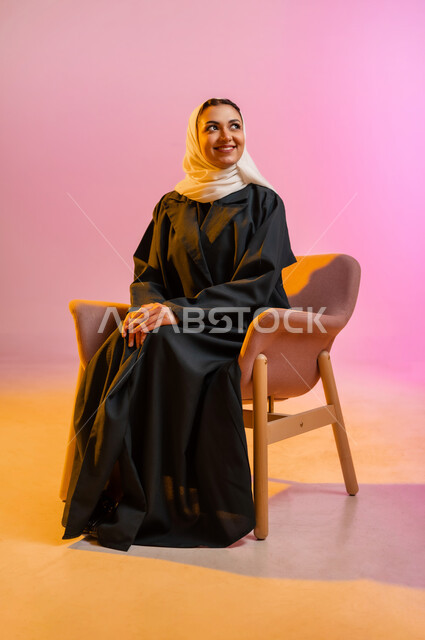مفهوم الانوثه والاهتمام بالمظهر الخارجي، تصاميم أزياء عصريه حديثه للمحجبات، بورتريه لسيده عربية خليجية عمانية ترتدي العباءة الملونه والحجاب تجلس على كرسي مريح بوضعيه التصوير والدعايه تنظر الى الاعلى بابتسامه، التسويق لأزياء المرأه المحجبه، خلفية ملونه