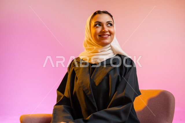 مفهوم الانوثه والاهتمام بالمظهر الخارجي، تصاميم أزياء عصريه حديثه للمحجبات، بورتريه لسيده عربية خليجية عمانية ترتدي العباءة الملونه والحجاب تجلس على كرسي مريح بوضعيه التصوير والدعايه تنظر الى الاعلى بابتسامه، التسويق لأزياء المرأه المحجبه، خلفية ملونه