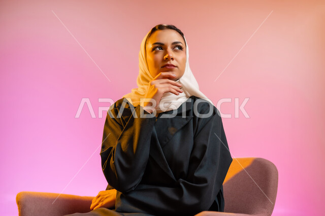 مفهوم الانوثه والاهتمام بالمظهر الخارجي، تصاميم أزياء عصريه حديثه للمحجبات، بورتريه لسيده عربية خليجية عمانية ترتدي العباءة الملونه والحجاب تجلس على كرسي مريح بوضعيه التصوير والدعايه تنظر الى الاعلى، التسويق لأزياء المرأه المحجبه، خلفية ملونه