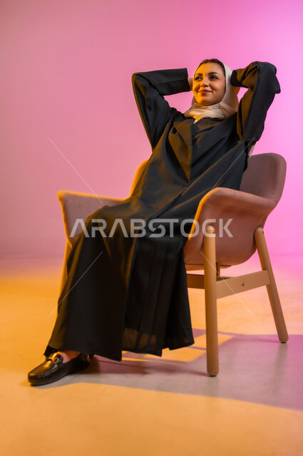 مفهوم الانوثه والاهتمام بالمظهر الخارجي، تصاميم أزياء عصريه حديثه للمحجبات، بورتريه لسيده عربية خليجية عمانية ترتدي العباءة الملونه والحجاب تجلس على كرسي مريح بوضعيه الاسترخاء تنظر الى الأعلى بابتسامه ، التسويق لأزياء المرأه المحجبه، خلفية ملونه