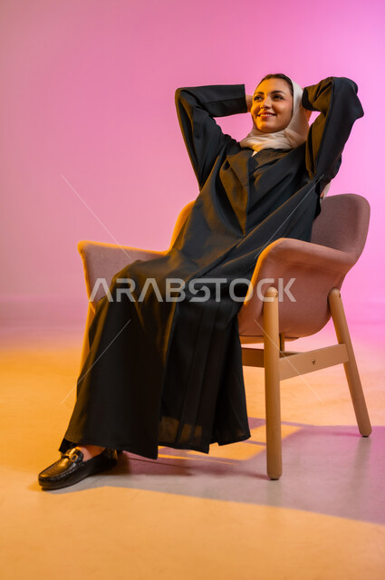 مفهوم الانوثه والاهتمام بالمظهر الخارجي، تصاميم أزياء عصريه حديثه للمحجبات، بورتريه لسيده عربية خليجية عمانية ترتدي العباءة الملونه والحجاب تجلس على كرسي مريح بوضعيه الاسترخاء تنظر الى الأعلى بابتسامه ، التسويق لأزياء المرأه المحجبه، خلفية ملونه