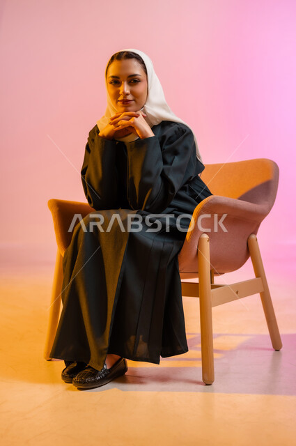 The concept of femininity and interest in external appearance, modern contemporary fashion designs for veiled women, a portrait of an Arab Gulf Omani woman wearing a colorful abaya and veil sitting on a comfortable chair in a photography and advertising position looking at the camera with a smile, marketing for veiled women’s fashion, colorful background