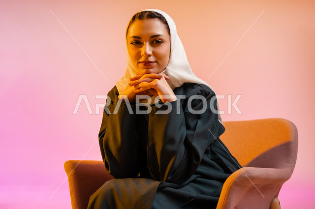 مفهوم الانوثه والاهتمام بالمظهر الخارجي، تصاميم أزياء عصريه حديثه للمحجبات، بورتريه لسيده عربية خليجية عمانية ترتدي العباءة الملونه والحجاب تجلس على كرسي مريح بوضعيه التصوير والدعايه تنظر الى الكاميرا بابتسامه، التسويق لأزياء المرأه المحجبه، خلفية ملونه