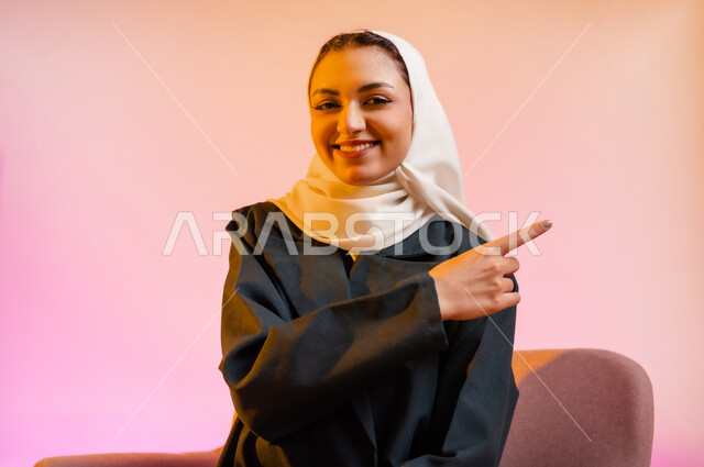 Sitting on a comfortable chair and pointing to a place, portrait of an Arab Gulf Omani woman wearing a colorful abaya and hijab sitting on a chair and pointing with her index finger in a direction with gestures of happiness and joy, movements indicating this place (this location), colorful background
