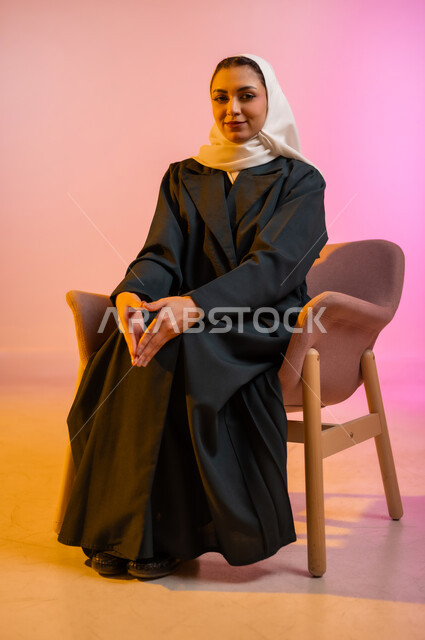 مفهوم الانوثه والاهتمام بالمظهر الخارجي، تصاميم أزياء عصريه حديثه للمحجبات، بورتريه لسيده عربية خليجية عمانية ترتدي العباءة الملونه والحجاب تجلس على كرسي مريح بوضعيه التصوير والدعايه تنظر الى الكاميرا بابتسامه، التسويق لأزياء المرأه المحجبه، خلفية ملونه