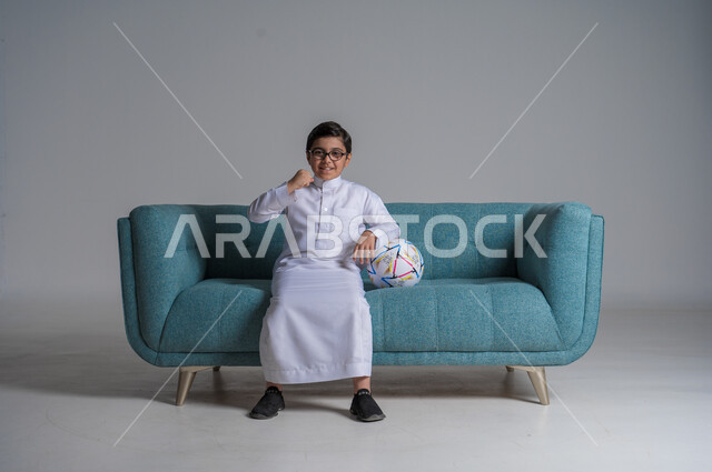 انشطة ترفيهية مسلية, قضاء أوقات الفراغ بالألعاب الحركية, بورتريه لفتى عربي خليجي سعودي يرتدي الثوب التقليدي  والنظارة الطبية يجلس على كنبه مريحة ويحمل بيده كرة القدم, خلفية رمادية