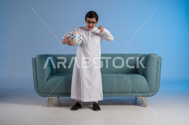 Having fun with favorite hobbies, passion for practicing motor sports, portrait of a Saudi Arabian Gulf boy wearing traditional dress and glasses holding a football with gestures of pleasure, full body image, gray background