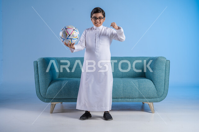الشغف بممارسة الرياضات الحركية, قضاء الوقت الممتع بالهوايات الترفيهية المفضلة, بورتريه لفتى عربي خليجي سعودي يرتدي الثوب التقليدي والنظارة الطبية يحمل بيده كرة القدم بإيماءات السرور, صورة الجسم بالطول الكامل, خلفية زرقاء