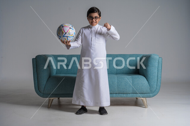 الشغف بممارسة الرياضات الحركية, قضاء الوقت الممتع بالهوايات الترفيهية المفضلة, بورتريه لفتى عربي خليجي سعودي يرتدي الثوب التقليدي والنظارة الطبية يحمل بيده كرة القدم بإيماءات السرور, صورة الجسم بالطول الكامل, خلفية رمادية