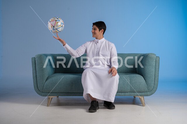 ممارسة الهواية المفضلة, شغف متابعة الأنشطة الرياضية, التسلية والترفيه بالرياضات الحركية, بورتريه لشاب عربي خليجي سعودي يرتدي الثوب التقليدي يجلس على كنبه مريحة ويحمل بيده كرة القدم بإيماءات السرور والسعادة, خلفية زرقاء