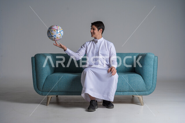 ممارسة الهواية المفضلة, شغف متابعة الأنشطة الرياضية, التسلية والترفيه بالرياضات الحركية, بورتريه لشاب عربي خليجي سعودي يرتدي الثوب التقليدي يجلس على كنبه مريحة ويحمل بيده كرة القدم بإيماءات السرور والسعادة, خلفية رمادية