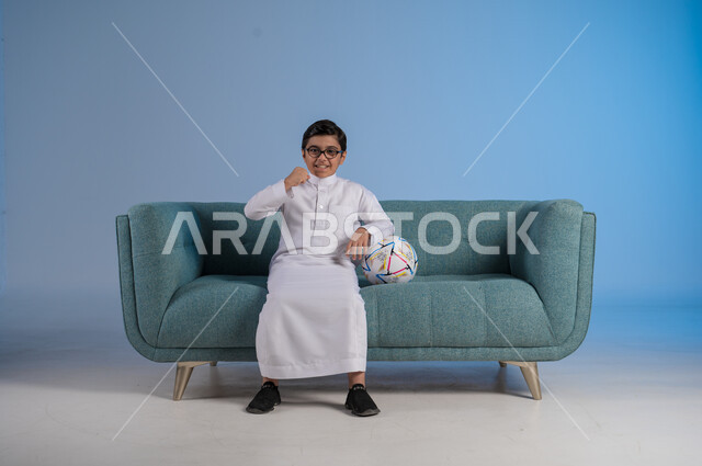 انشطة ترفيهية مسلية, قضاء أوقات الفراغ بالألعاب الحركية, بورتريه لفتى عربي خليجي سعودي يرتدي الثوب التقليدي والنظارة الطبية يجلس على كنبه مريحة ويحمل بيده كرة القدم, خلفية رمادية